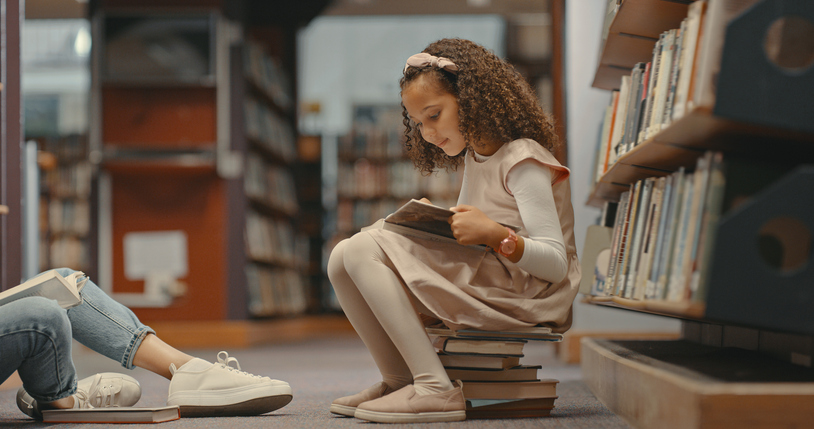 Young girl siiting on books inside the library with unrecognizable person. Girl with curly hair reading a book. Mixed race child doing research