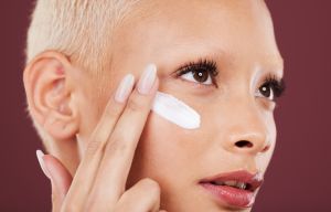 Face, skincare and lotion with a model black woman in studio on a red background to apply moisturizer. Facial, beauty or cream with an attractive young female applying antiaging treatment to her face