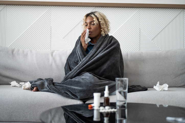 Sick young black woman sitting under the blanket on sofa and sneeze with tissue paper at home. Female blowing nose, coughing or sneezing in tissue at home, suffering from flu. Cold and fever concept