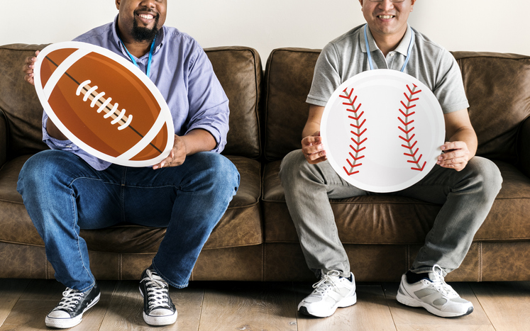 Men holding baseball and rugby icons sitting on couch