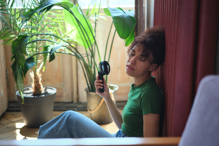 Summer heat. African American woman using portable fan at home surviving a heatwave, sitting by window enjoying fresh air, cooling down on hot summer day indoors